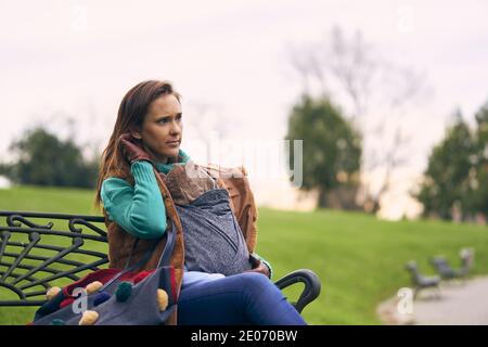 Schöne Frau im Pullover Mantel sitzen auf einer Bank im Winter tragen ihren Sohn an einem bewölkten Tag. Mutterschaft Lifestyle-Konzept Stockfoto