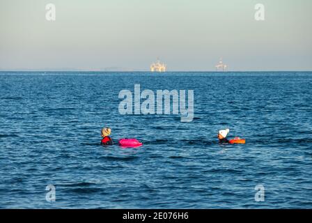 Zwei Menschen schwimmen in der Nordsee mit Ölbohrinseln im Hintergrund, in Neoprenanzügen, Kirkcaldy, Fife, Schottland Stockfoto