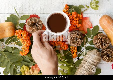 Florale Herbstkomposition. Eine Tasse Kaffee in der Hand einer Frau auf weißem Holzhintergrund mit gelb fallenden Blättern Ahorn und Kegel. Hallo Herbst. Flach Stockfoto