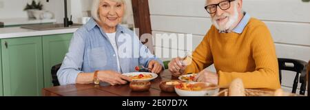 Lächelndes, älteres Paar, das auf die Kamera schaut, während es am Tisch sitzt und auf verschwommenem Hintergrund ein vegetarisches Abendessen mit Transparent serviert Stockfoto