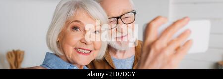 Fröhliche ältere Frau, die Kamera anschaut, während sie Selfie mit Mann auf verschwommenem Vordergrund drinnen macht, Banner Stockfoto