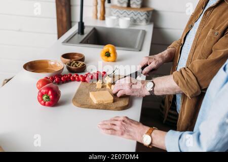 Beschnittene Ansicht der älteren Frau in der Nähe des Ehemanns, der Käse anschneidet Schneidebrett in der Küche auf unscharfem Hintergrund Stockfoto