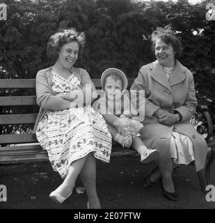 1950er Jahre, historische, glückliche Familienzeiten, eine Mutter, die auf einer Bank sitzt und zusammen mit ihrer Tochter lacht, die eine Mütze trägt und deren Hand in einem knusprigen Paket ist, zusammen mit der Großmutter des Kindes, die ein großes Lächeln im Gesicht hatte, England, Großbritannien. Stockfoto