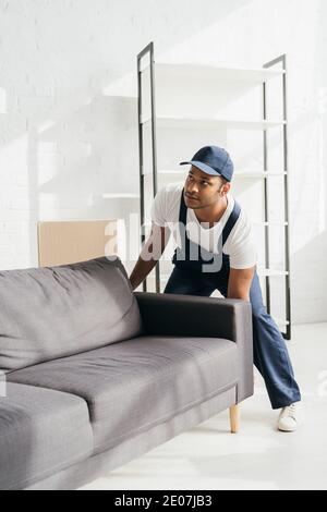 Junge indische Mover in Uniform und Mütze tragen Sofa in Wohnung Stockfoto