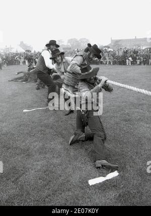 Siebziger Jahre, historisch, draußen auf einem Dorffest, ein Team von erwachsenen Männern in Cowboy-Outfits ziehen an einem Seil, während sie in einem Tauziehen-Krieg-Wettbewerb, England, Großbritannien konkurrieren. Stockfoto