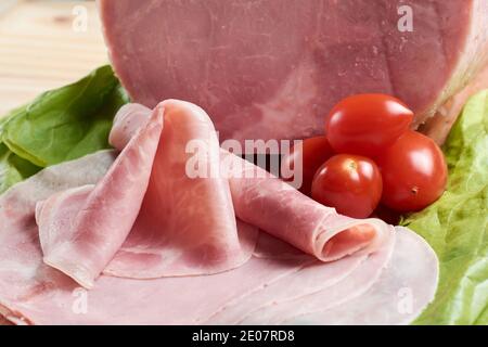Köstlicher, in Scheiben geschnittener und gerollter gekochter Schweineschinken auf einem Holztisch, begleitet von frischen Kirschtomaten und Salat. Stockfoto