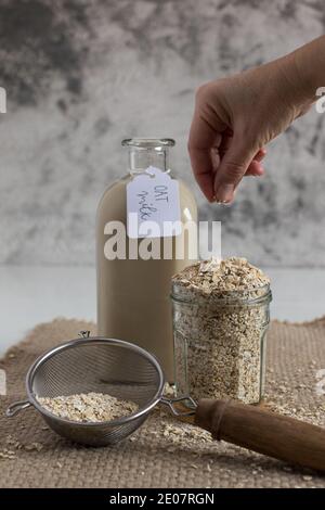 Frau, die Haferstücke in ein Glas voll mit diesem Müsli neben einer gekennzeichneten Hafermilchflasche von Hand fallen lässt. Vertikale Ansicht. Stockfoto