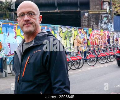 Ein Mann mit kleinen runden Gläsern läuft vor Santander Bikes Docking Station & Graffiti Wand auf Brick Lane, Shoreditch, East London. Stockfoto