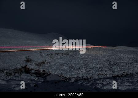 Glencoe, Schottland, Großbritannien. Dezember 2020. Im Bild: Die A82 mit einem einsamen Fahrzeug in der Nacht unter dem abnehmenden Herbstmond gesehen. Der Schnee reflektiert das Licht zurück und verursacht ein dramatisches Nachtbild. Gelbe Schneewarnung an Ort und Stelle, da mehr Schnee mit frostigen Temperaturen über Nacht erwartet wird. Quelle: Colin Fisher/Alamy Live News Stockfoto