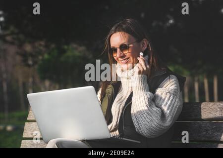 Frau in Kopfhörer arbeitet mit Laptop draußen im Park. Freiberufliche Arbeit. Online arbeiten oder studieren. Neues technologisches Nutzungskonzept Stockfoto