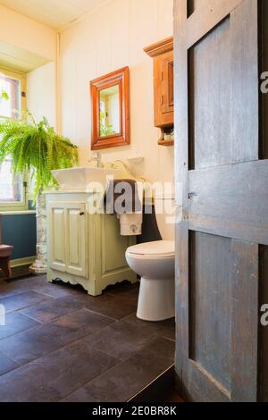 Offene Tür des Hauptbadezimmers mit keramischen Fliesenboden und Weiße Toilette und Waschbecken auf dem antiken Schrank innen Ein altes Haus im Cottage-Stil von 1841 Stockfoto