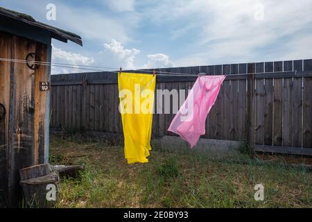 Saubere Kleidung wird an einem Seil getrocknet. Zwei Regenmäntel in rosa und gelben Farben flattern im Wind im Hof vor dem Hintergrund des Zauns. Stockfoto