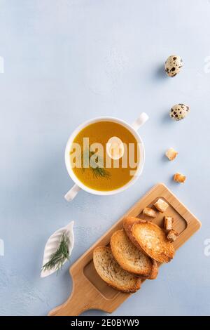 Bouillon in weißer runder Suppenschüssel, Wachtelei und Fenchelblatt auf blauem Hintergrund. Geröstetes Brot auf Holzbrett. Flach liegend. Vertikale Ausrichtung. Homema Stockfoto