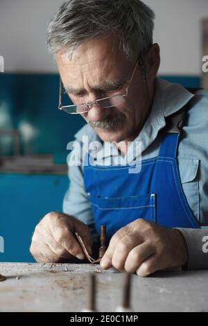 Ein älterer Meister mit Brille bei der Arbeit. Ein weißer alter Mann mit kaukasischem Aussehen sitzt an einem Tisch und arbeitet mit seinen Händen Stockfoto