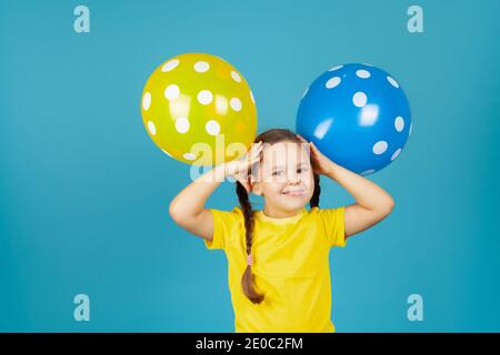 Nahaufnahme lachendes Mädchen mit Zöpfen in einem gelben T-Shirt hält Luftballons wie Ohren an ihrem Kopf, isoliert auf blauem Hintergrund Stockfoto