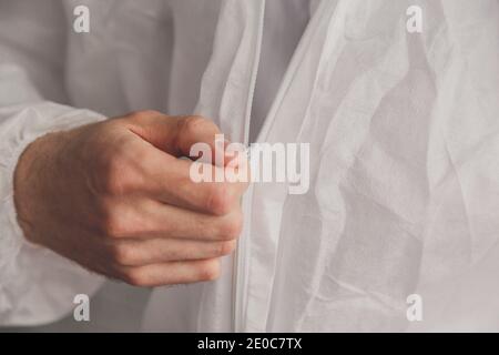 Der Arzt epidemiologist legt den weißen Schutzüberanzug an, befestigt den Reißverschluss, die Hände aus der Nähe. Hände in Gummischutzhandschuhe. Persönlicher Schutz Stockfoto