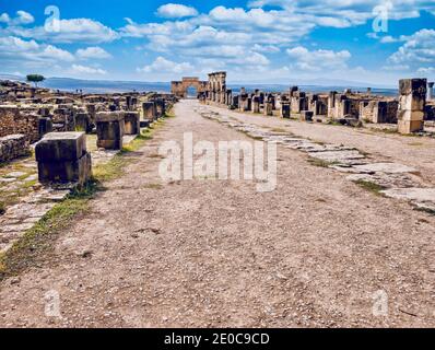 Eine Straße, die zu einem Triumphbogen aus dem 3. Jahrhundert führt, führt durch die Ruinen der antiken römischen Stadt Volubilis in Marokko. Stockfoto
