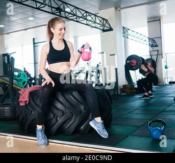 Fröhliche junge Frau in Sportbekleidung hebt die Kettlebell mit einer Hand auf dem großen Rad für funktionelles Training. Stockfoto