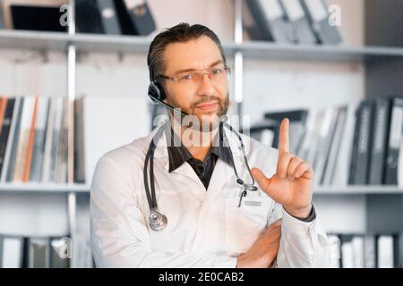 Arzt mit Headset gibt online Ratschläge. Er hört aufmerksam zu und lenkt mit Gesten die Aufmerksamkeit auf den Patienten. Stockfoto