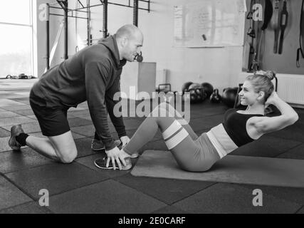 Der Mann hält die Beine der Frau, die die Verdrehung Übung für die Bauchmuskeln auf der Matte in der Turnhalle liegt. Stockfoto