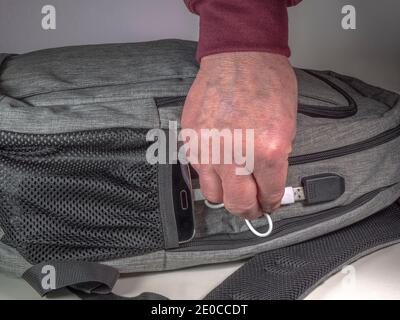 Man’s Hand verbindet ein Kabel / Kabel zwischen einem Handy in einer Tasche auf einem Rucksack / Rucksack, und ein USB-Ladegerät Anschluss in der Tasche angebracht. Stockfoto
