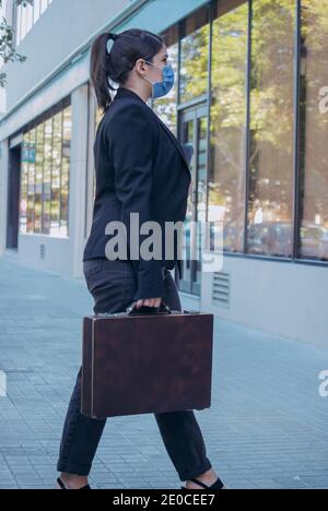 Elegante Geschäftsfrau, die ihre Maske trägt, auf dem Weg zum Eingang ihrer Büros, um ihren Arbeitstag früh am Morgen zu beginnen. Mädchenboss 2021. Stockfoto