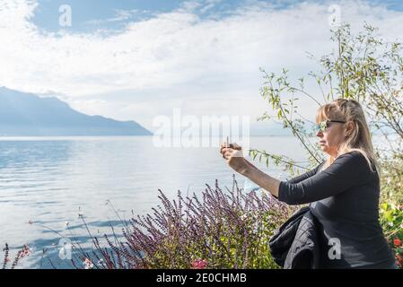 Blonde Frau, die ein Foto mit dem Handy in einem See, umgeben von Bergen und Blumen. Montreux, Genfersee in der Schweiz Stockfoto