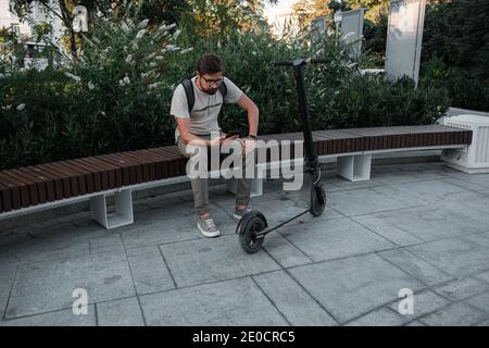 Hipster Mann Pendler mit Elektroroller im Freien in der Stadt, mit Smartphone. Junge Millennial Kerl mit Handy-Verleih von Öko-Transport in su Stockfoto