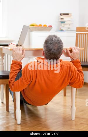 Reifer Mann tut Pull-ups zu Hause unter einem Esstisch Mit Laptop - konzentrieren Sie sich auf den Kopf Stockfoto