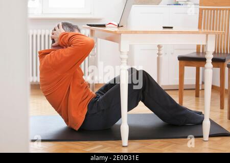 Reifer Mann tut Sit-ups zu Hause unter einem Esstisch Mit Laptop und Smartphone - konzentrieren Sie sich auf den Mann Stockfoto