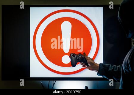 Mann in der Haube, der den Controller vor dem Bildschirm hält und eine rote Ausrufemeldung anzeigt, während ein Spiel aufgrund von Bugs, Hacks oder Problemen abstürzt Stockfoto