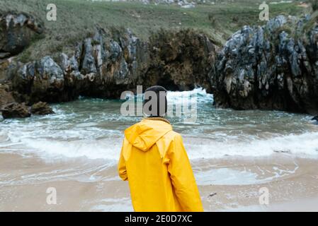 Rückansicht einer nicht erkennbaren weiblichen Forscherin in gelbem Regenmantel Entspannen Sie sich an der steinigen Küste des Cobijero Beach und bewundern Sie erstaunlich Blick auf den See auf dem rüber Stockfoto