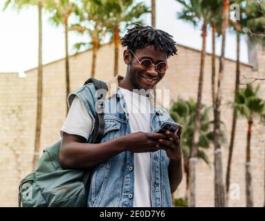 Niedriger Winkel des Inhalts junge afroamerikanische Hipster männlicher Reisender Mit Dreadlocks im trendigen Outfit und Sonnenbrillen per Handy Im Stehen ne Stockfoto