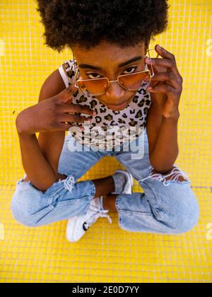 Von oben junge afroamerikanische Frau in trendigen Ripped Jeans Und stilvolle Sonnenbrille auf gelbem Boden Stockfoto