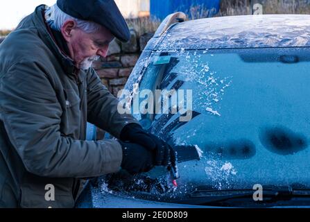 Senior man kratzt Eis von der Windschutzscheibe im Winter mit Frostmustern auf dem Autodach, Schottland, Großbritannien Stockfoto