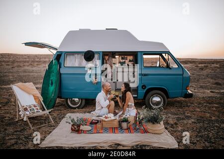 Seitenansicht eines Reisenden Paares, das auf einer Decke in der Nähe des Transporters sitzt In Savanna und mit Picknick während der Sommerferien genießen und Einander anschauend Stockfoto
