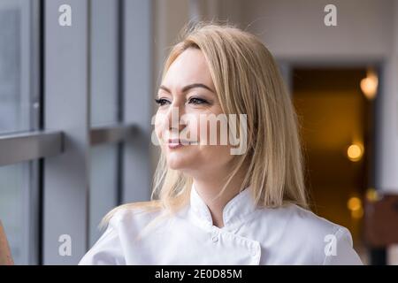 Lächelnde weibliche Sanitäter in weißer Uniform stehend mit den Händen in Taschen im Flur des Krankenhauses und Blick aus dem Fenster Stockfoto