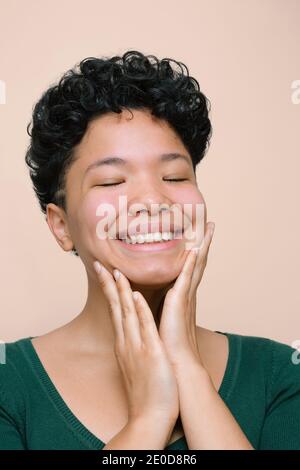 Die junge Latina lächelt und berührt ihr Gesicht mit den Händen. Geschlossene Augen. Isoliertes vertikales Foto, beigefarbener Hintergrund Stockfoto