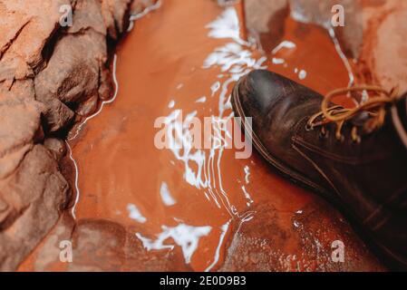 Von oben der Ernte unkenntlich Wandermann in Trekkingstiefeln In der Nähe von Pfütze mit orangefarbenem Wasser in Sandsteinschlucht stehend Wadi Rum Stockfoto