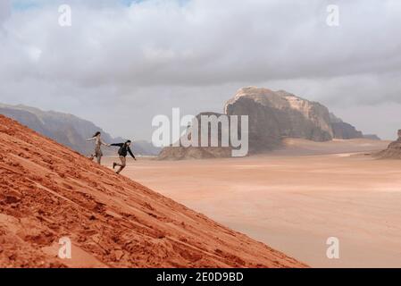 Seitenansicht von ein paar Reisenden, die den sandigen Hügel hinunterlaufen Im Wadi Rum Sandsteintal, während Sie Freiheit und Urlaub genießen In Jordanien Stockfoto