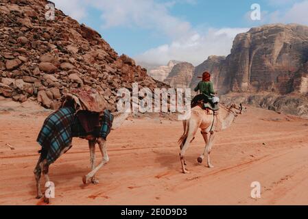 Rückansicht des Reisenden auf Kamel sitzen und entlang reiten Sandsteintal mit Felsen in Wadi Rum Stockfoto