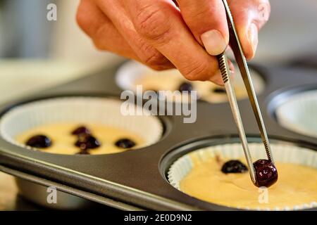 Nahaufnahme von Ernte anonymen Koch mit Küchenzange setzen Beeren Zu Teig in Form von Muffins während der Zubereitung von Gebäck in der Küche Stockfoto