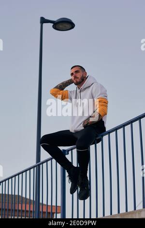 Von unten Vollkörper von erwachsenen bärtigen Männchen mit tätowiert Arme gekleidet in lässigen Hoodie sitzen auf Metallzaun in der Nähe Straßenlaterne in der Stadt Stockfoto