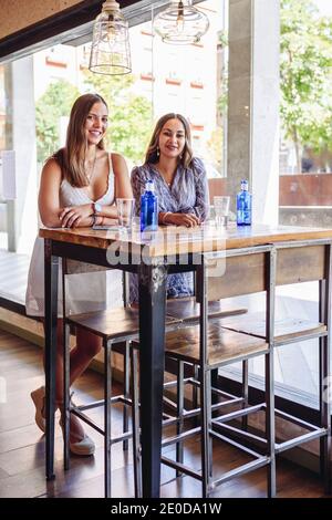 Lächelnde junge Freundinnen, die die Kamera betrachten, während sie am Tisch stehen Mit Getränken während der Tagung in der modernen Cafeteria am Sommertag Stockfoto