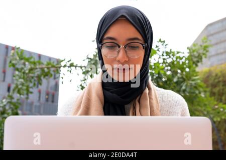Beschäftigte arabische Unternehmerin im traditionellen Kopftuch auf der Straße sitzen Mit Laptop und Arbeit an neuen Business-Projekt Stockfoto