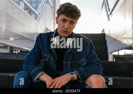 Junger Mann mit lockigen Haaren in Denim-Kleidung mit Wireless Kopfhörer um den Hals sitzen in Treppen in der Stadt suchen Bei der Kamera Stockfoto