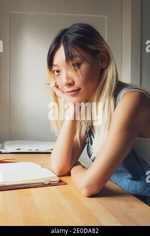 Seitenansicht der nachdenklichen asiatischen weiblich sitzen am Tisch mit Tagebuch und sich an der Hand lehnen, während man auf die Kamera schaut Stockfoto