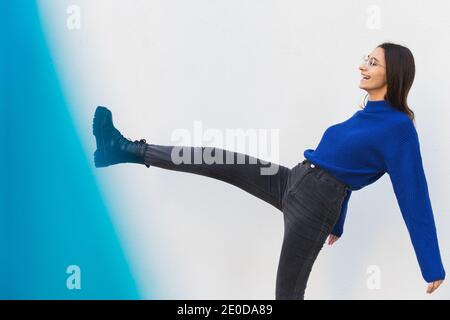 Seitenansicht der fröhlichen weiblichen Millennial im stilvollen Outfit stehen Auf der Straße mit erhobenem Bein und Spaß haben Stockfoto