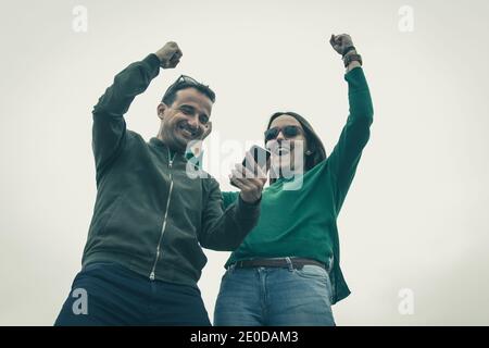 Euphorisches Paar mit Faust nach oben Blick auf Handy über weißem Hintergrund. Junge Frau und Mann feiern gute Nachrichten. Lotterie Gewinner Konzept Stockfoto