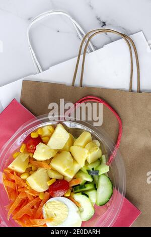 Draufsicht auf leckeren Salat mit verschiedenen Gemüse und gekocht Ei in Plastikschüssel mit Papiertüten auf Tisch gelegt Zum Mitnehmen gesunde Lebensmittel Lieferung Stockfoto